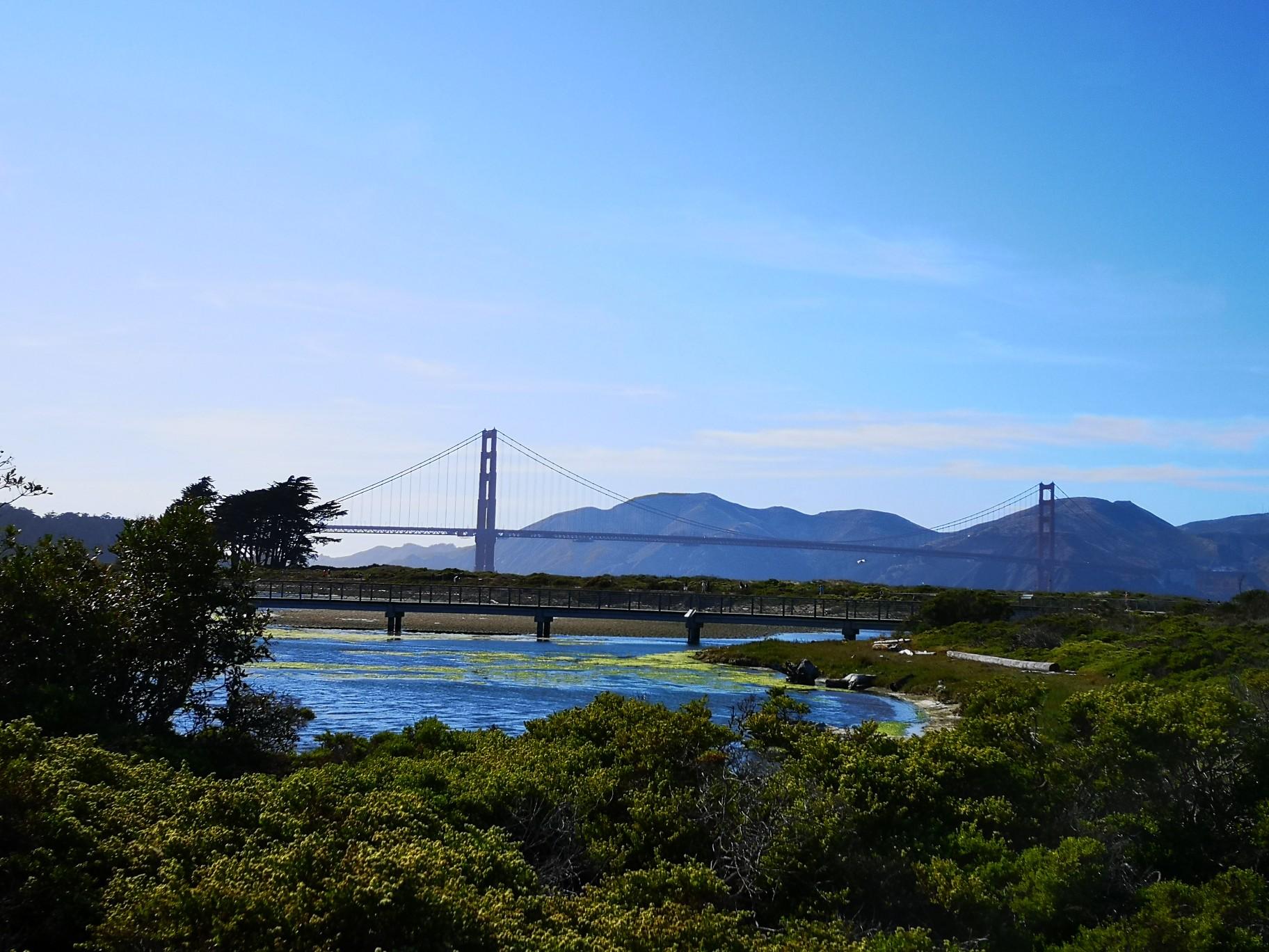 Iconic bridge spans water with distant mountains under a bright blue sky.