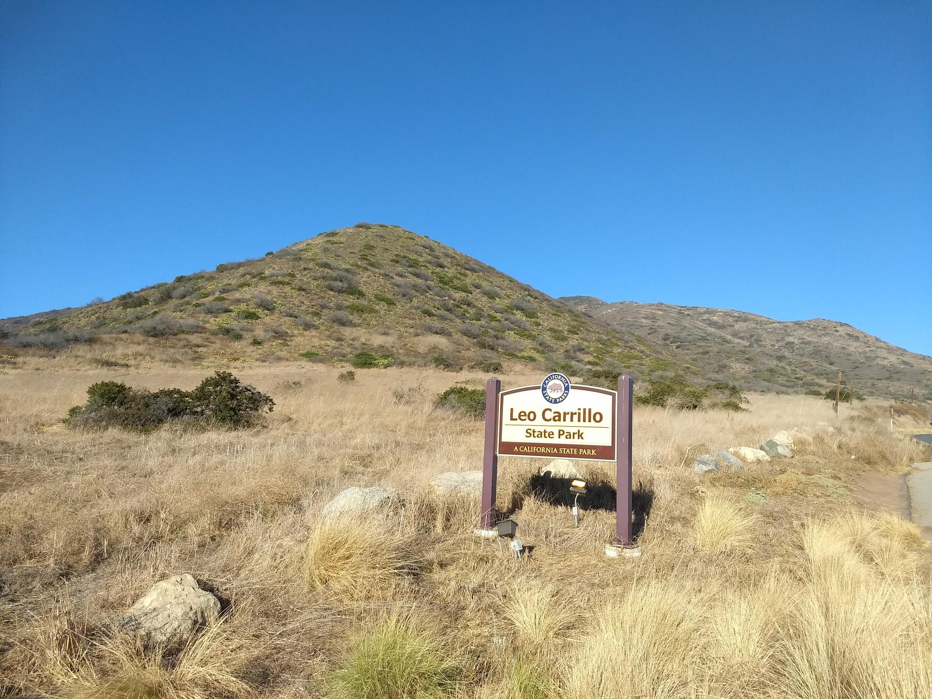 A wooden park sign stands in a dry, grassy field under a clear sky.
