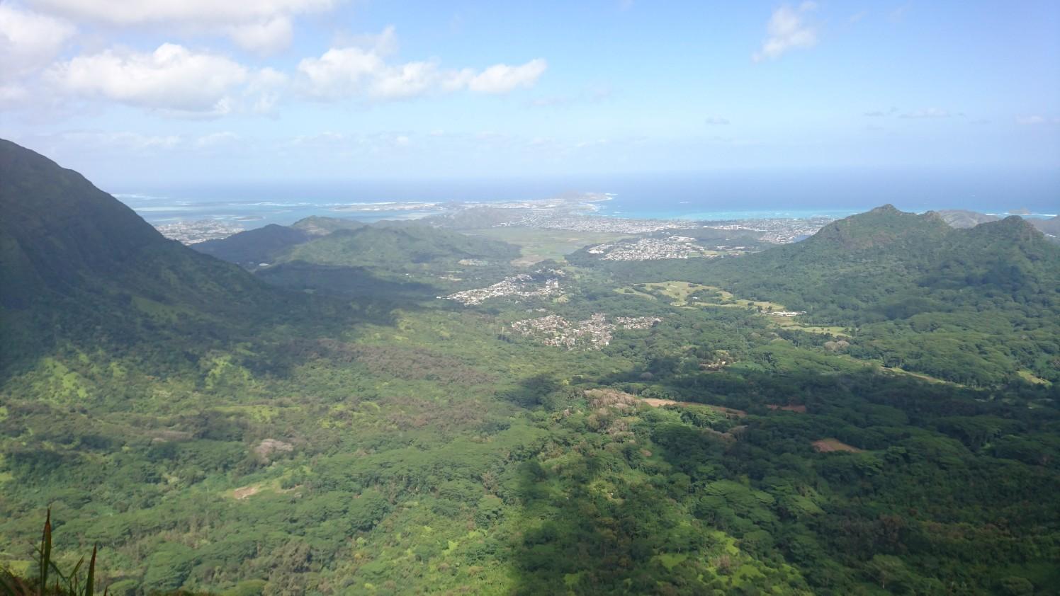 Wide view of green mountains, a valley with buildings, and a distant blue ocean.