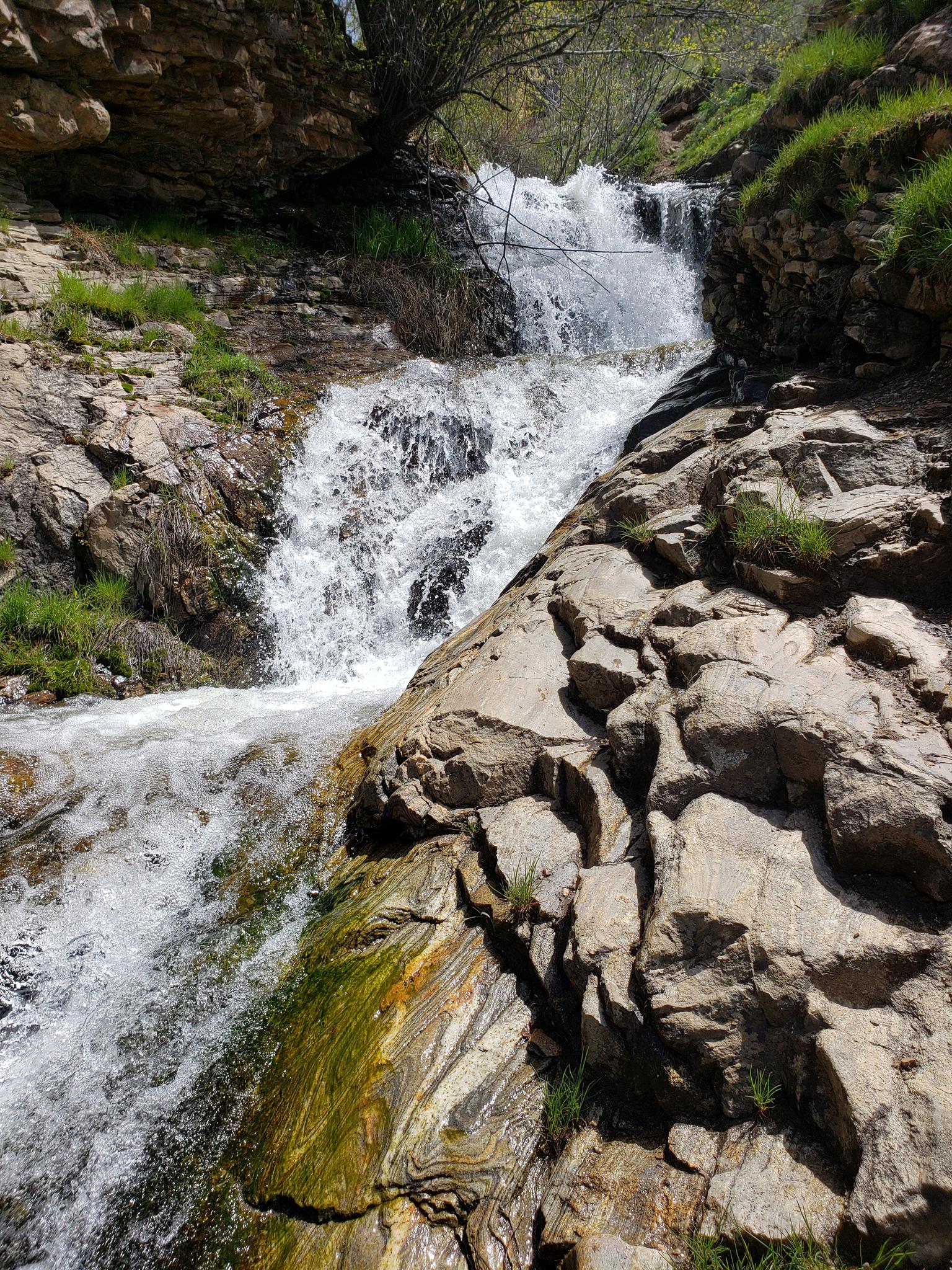 A multi-tiered waterfall cascades over mossy rocks in a sunny, green landscape.