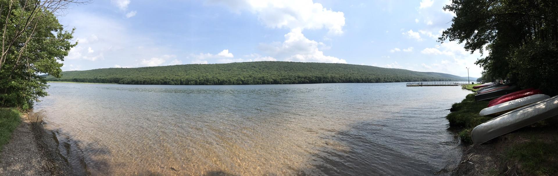 Panoramic view of a wide lake with forested hills and canoes on the shore.