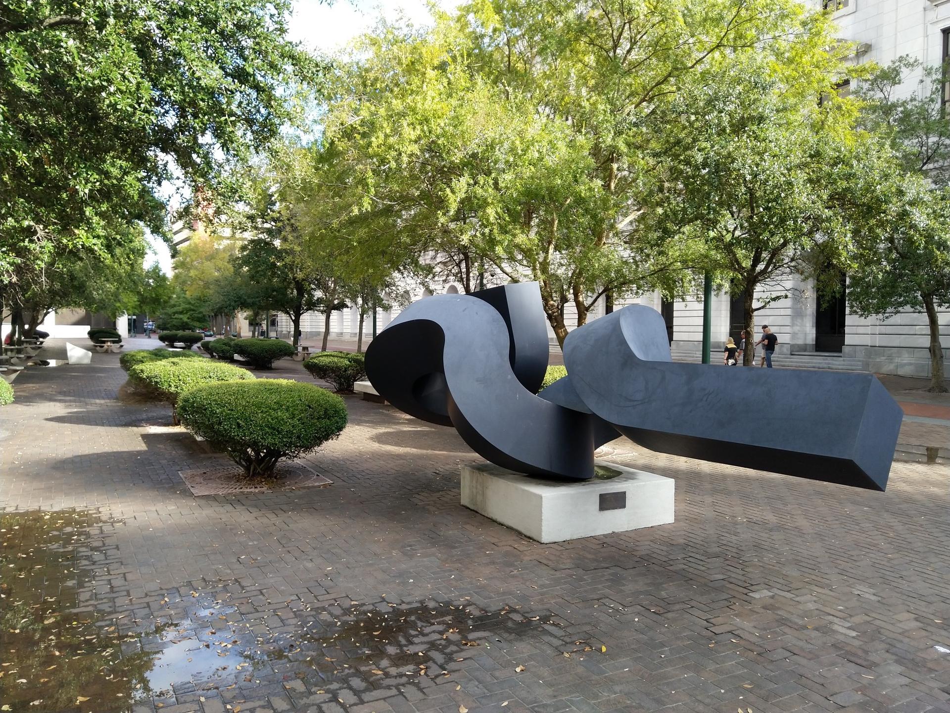 A large, dark abstract sculpture sits on a paved path in a green park.