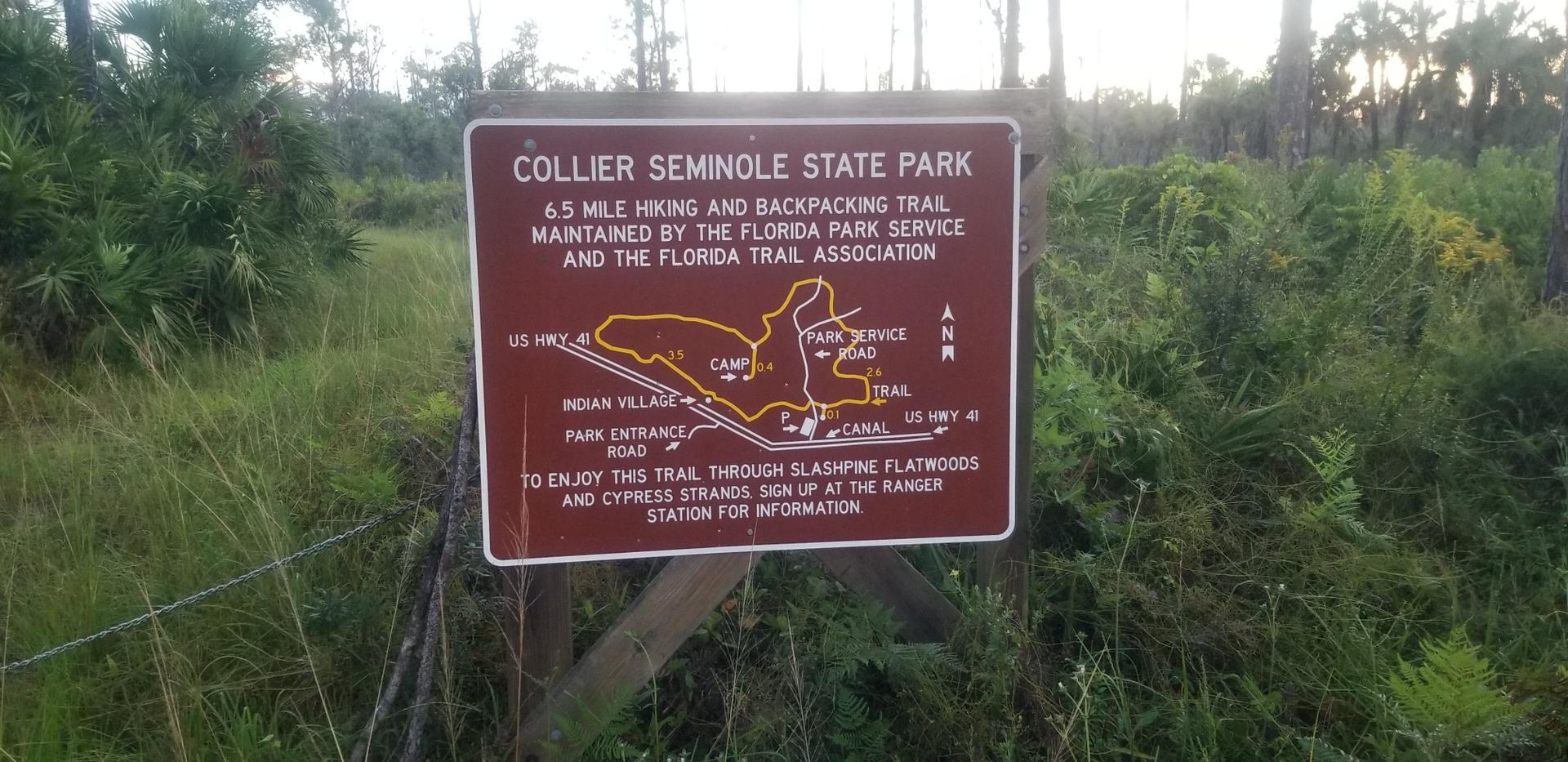 A brown park sign with a trail map stands amidst lush green foliage under natural light.