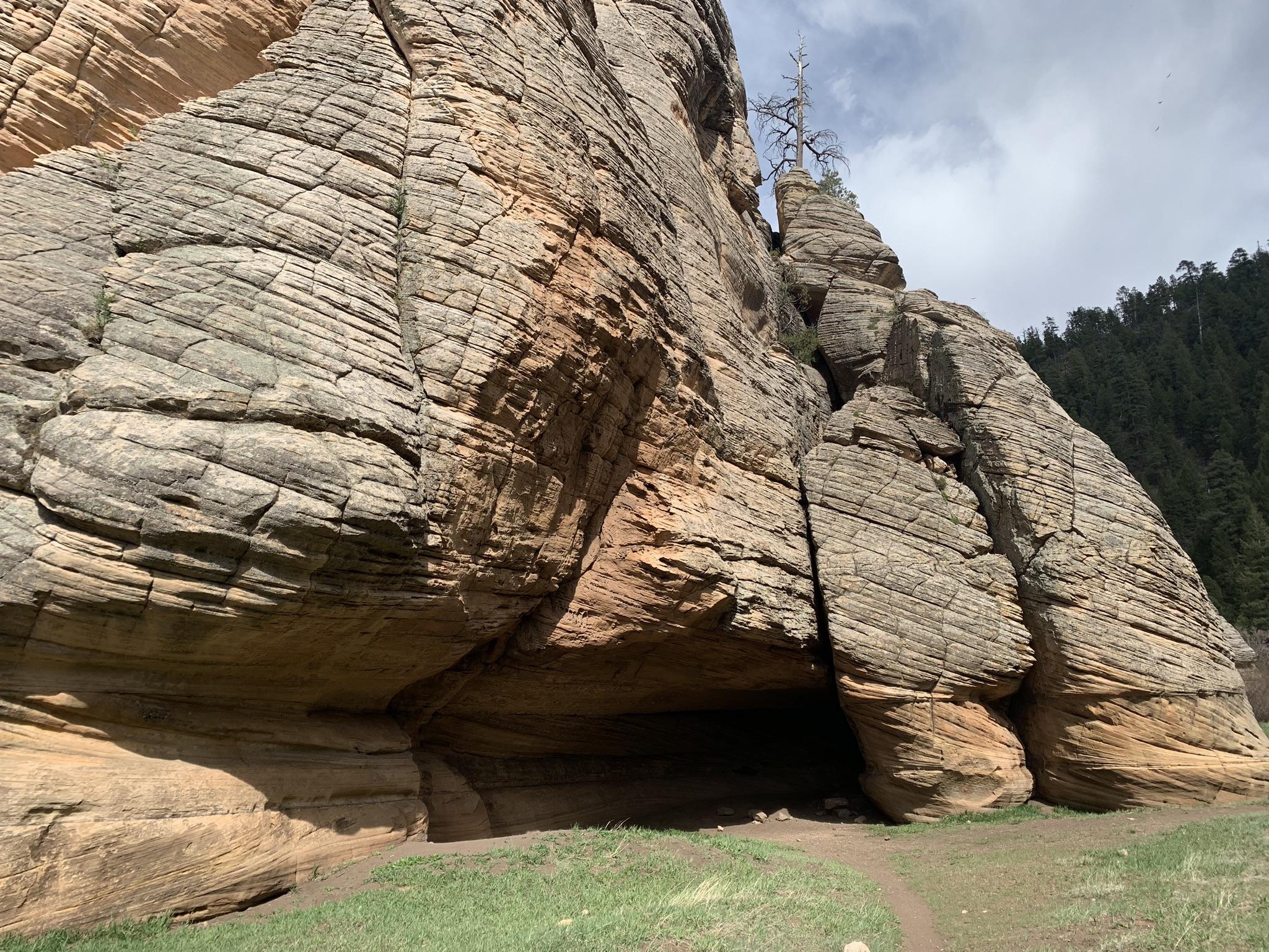 Massive layered rock formation with a dark cave opening under a cloudy sky.