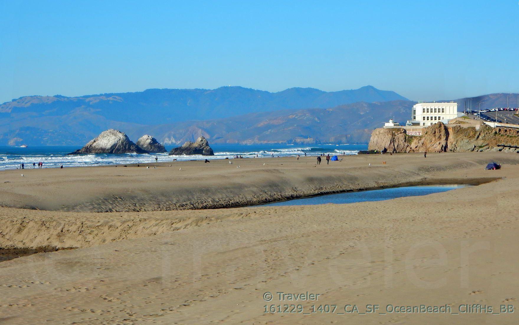 Sandy beach with ocean, rock formations, a building, and distant mountains.