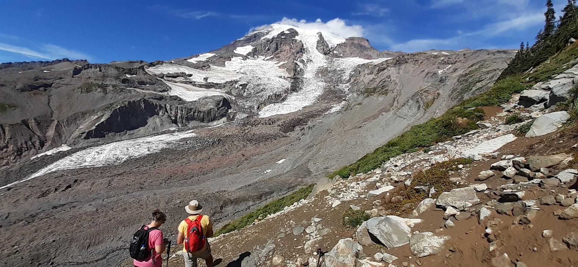 Two hikers on a rocky trail overlook a large snow-capped mountain and glacier.