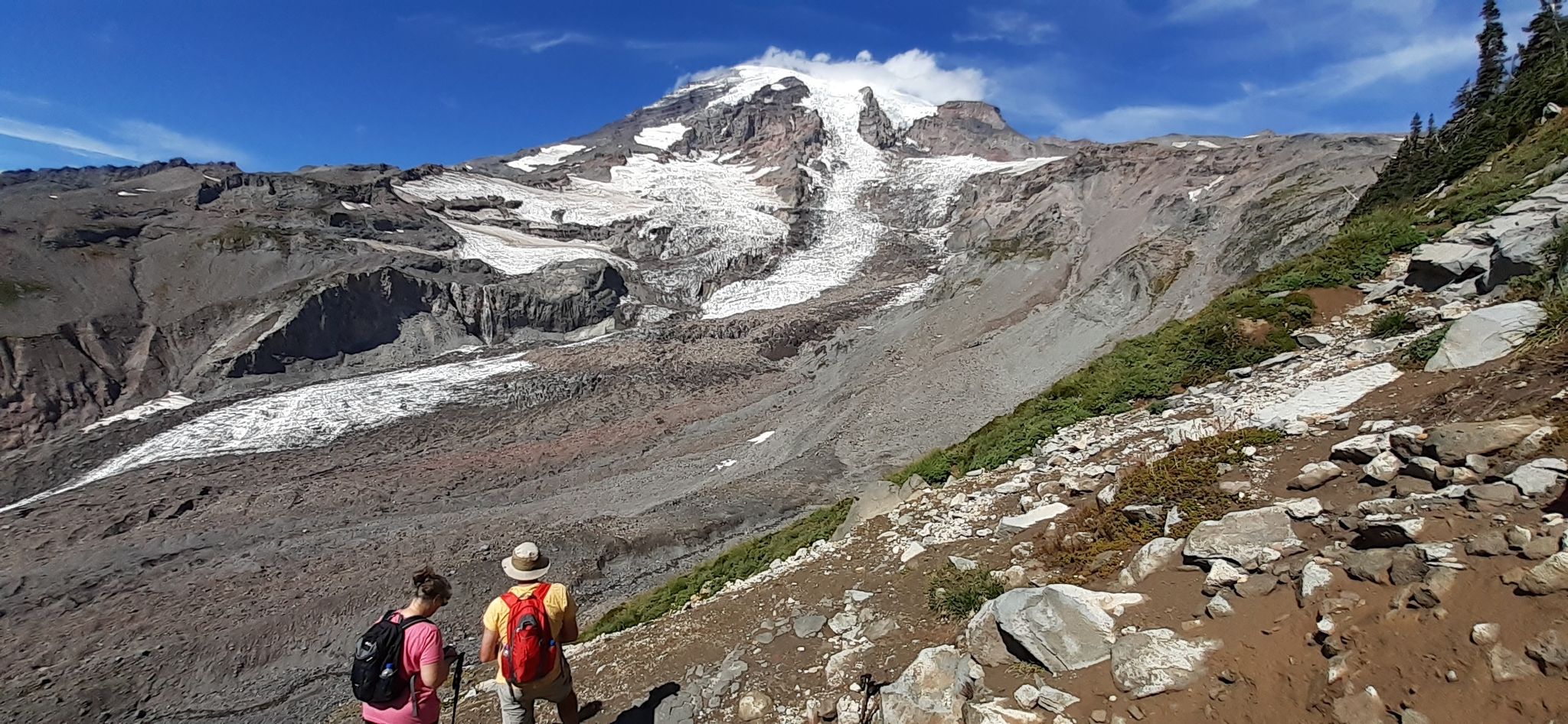 Two hikers on a rocky trail overlook a large snow-capped mountain and glacier. - Panorama Point – Mount Rainier National Park 