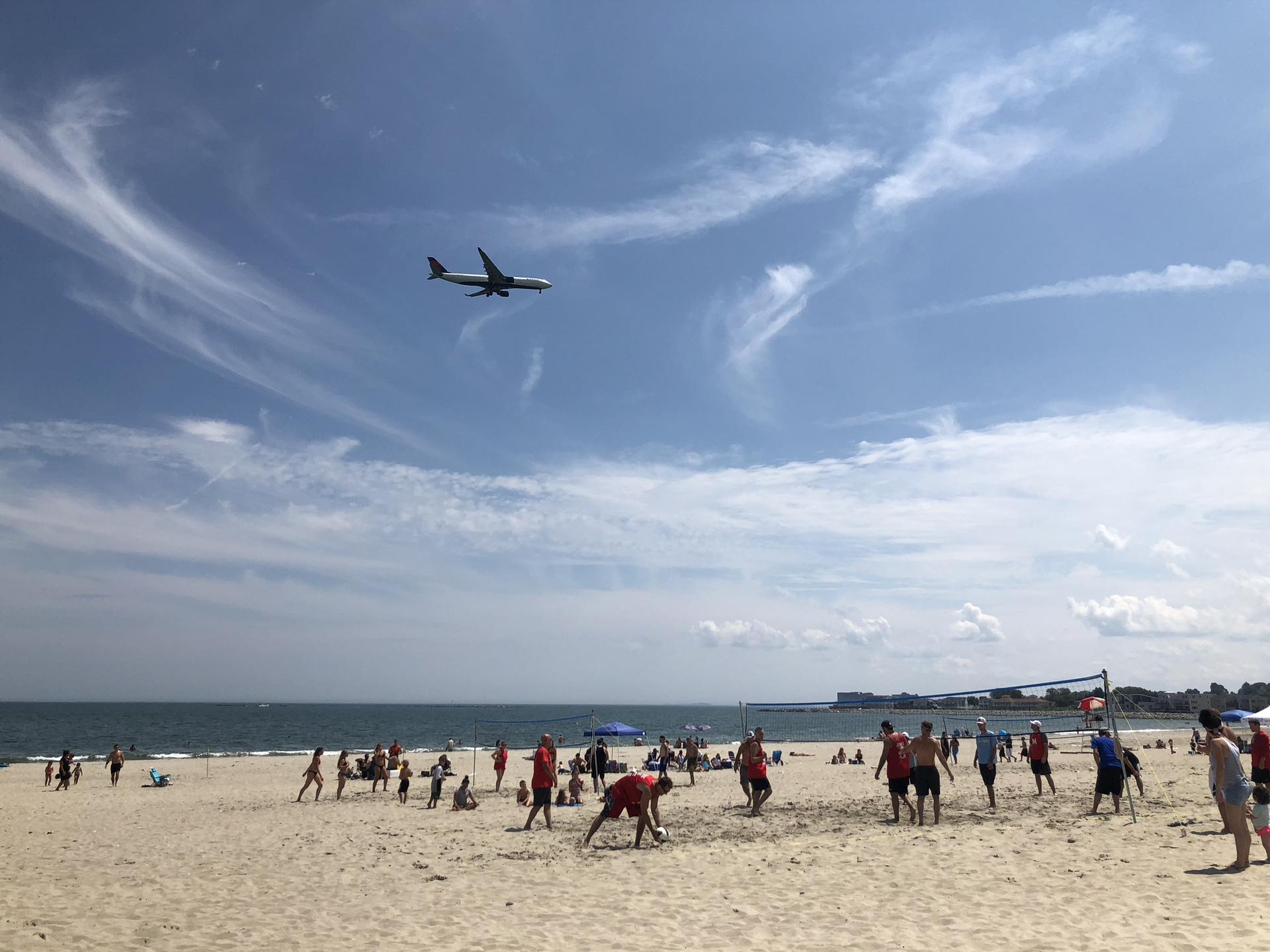 Crowded beach with people playing volleyball, airplane flying overhead.