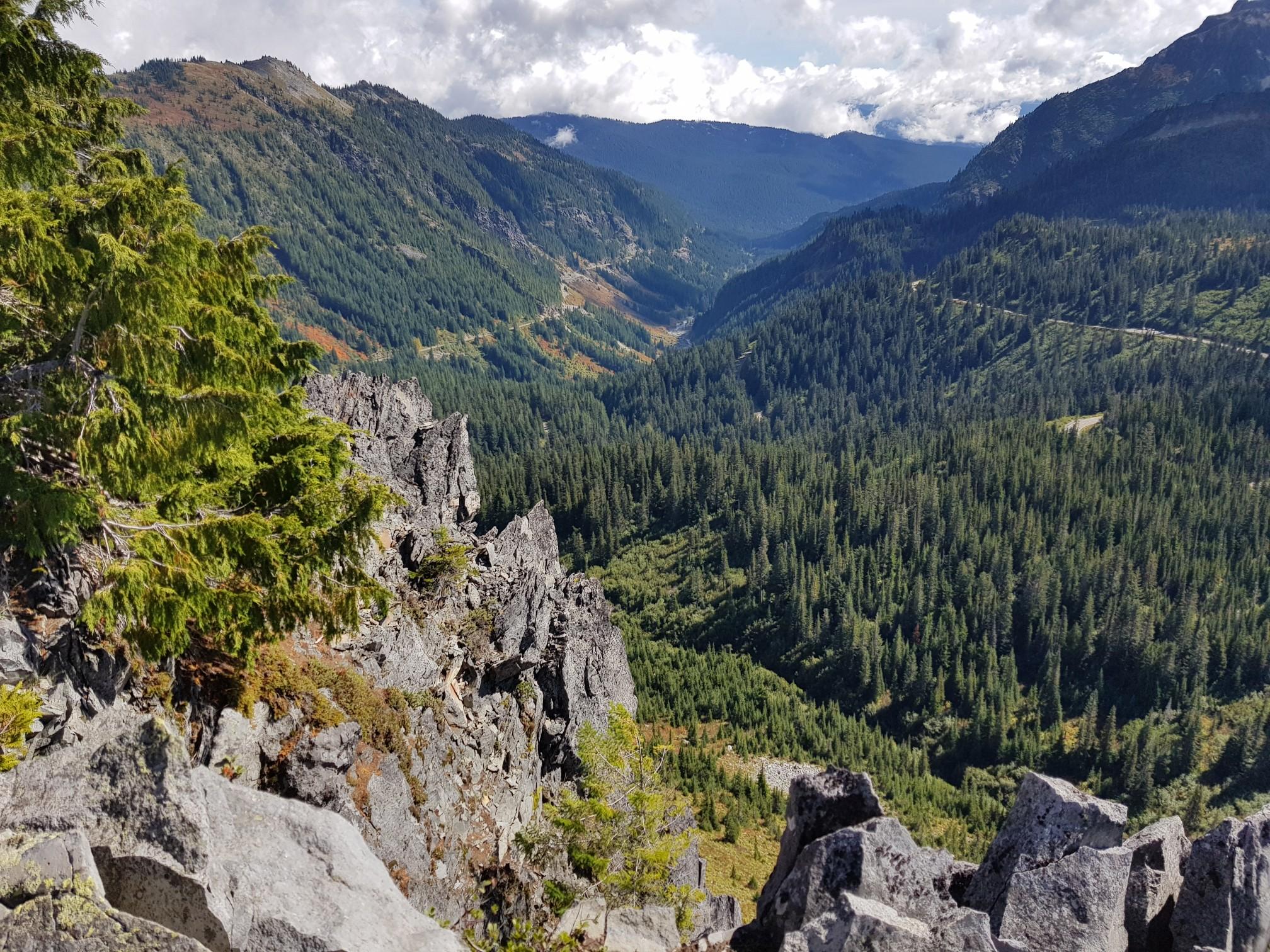 Rugged rocky peaks overlook a vast forested valley under a cloudy sky.