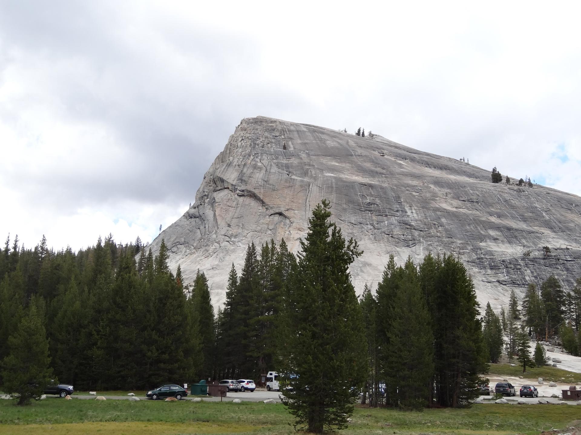 Large granite dome overlooks a pine forest and parking lot under cloudy skies.