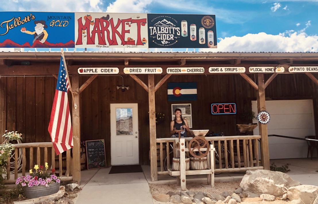 A person stands on the porch of a rustic market building with signs.