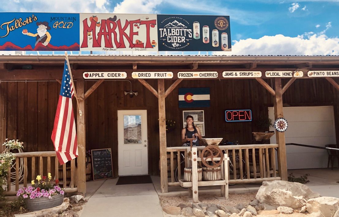 A person stands on the porch of a rustic market building with signs. - Fruit and Wine Byway - East Orchard Loop