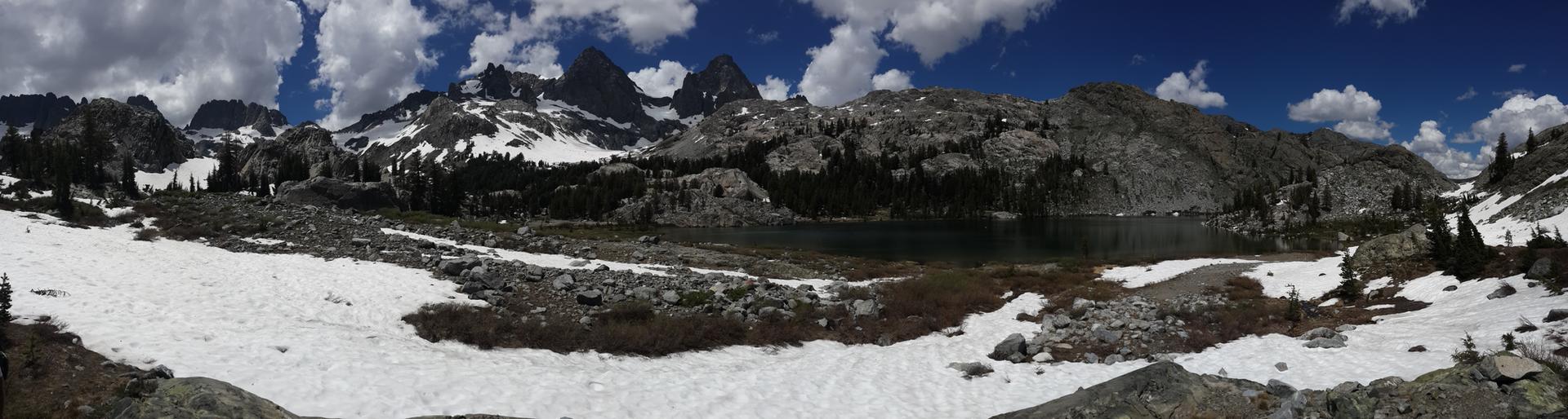 Panoramic mountain lake with snow-dusted peaks under a bright, partly cloudy sky.