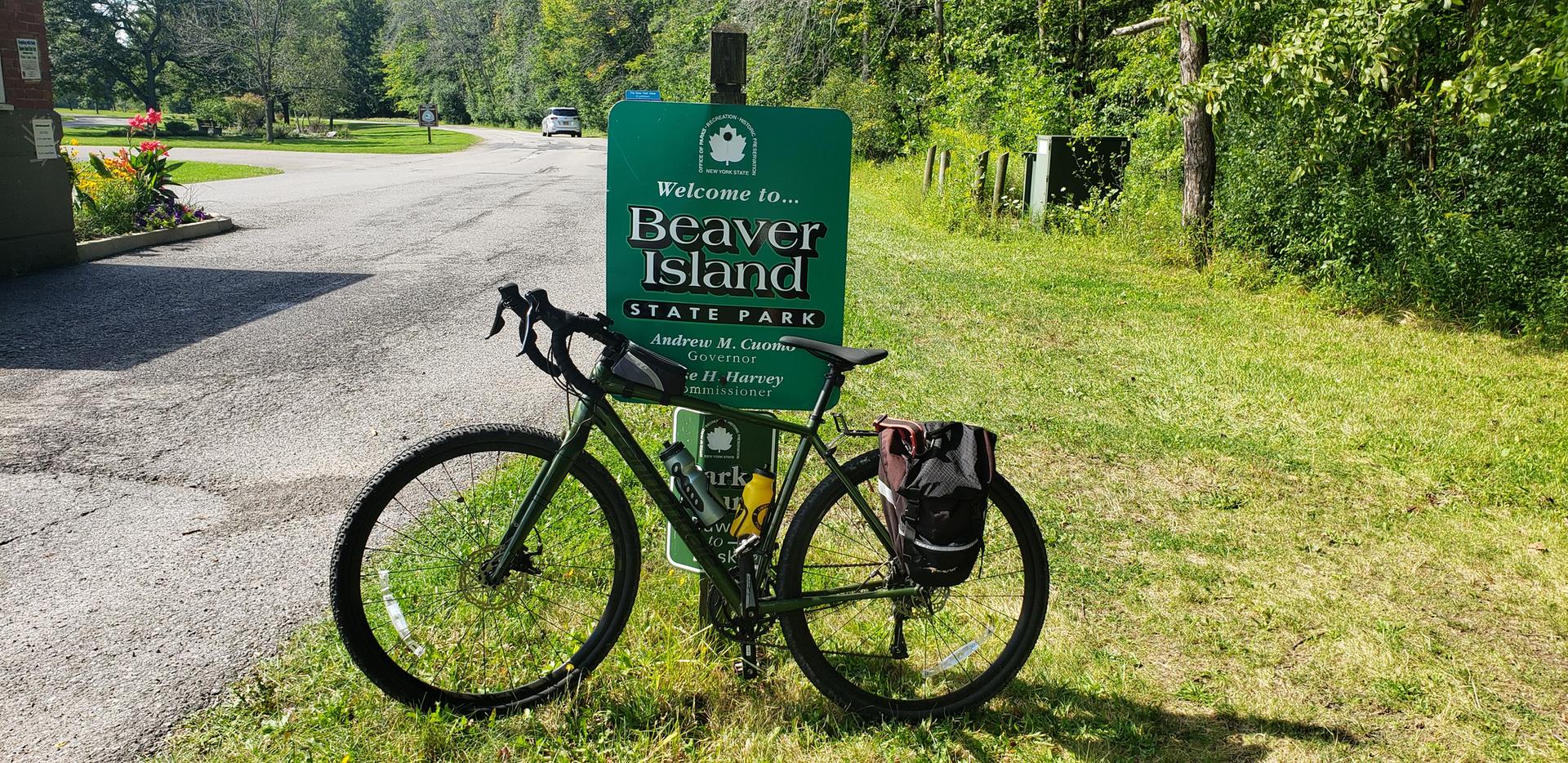 A touring bicycle with bags parked next to a welcome sign on a sunny day.