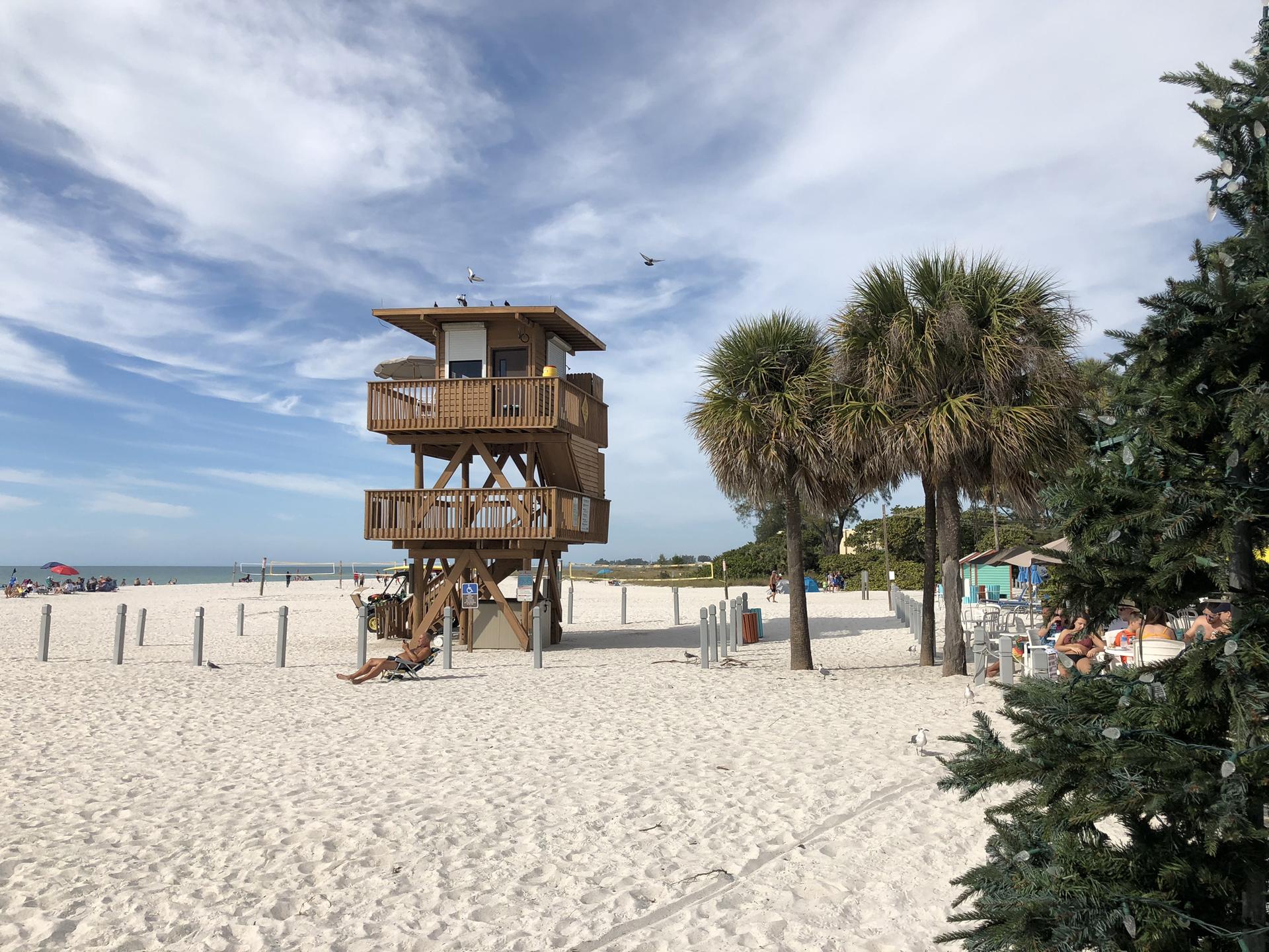 Lifeguard tower on a sunny sandy beach with palm trees and distant people.