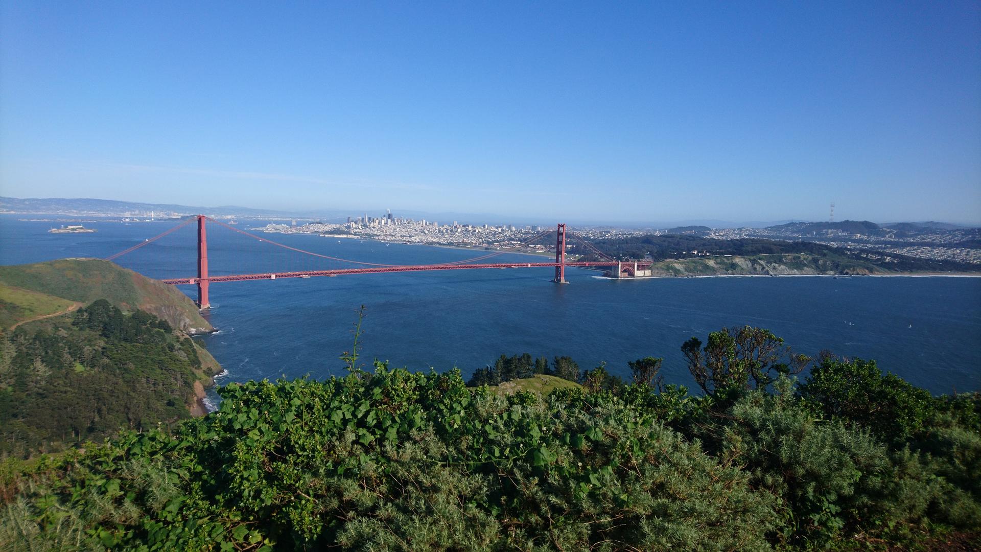 Wide view of a red suspension bridge over a bay, with a distant city.