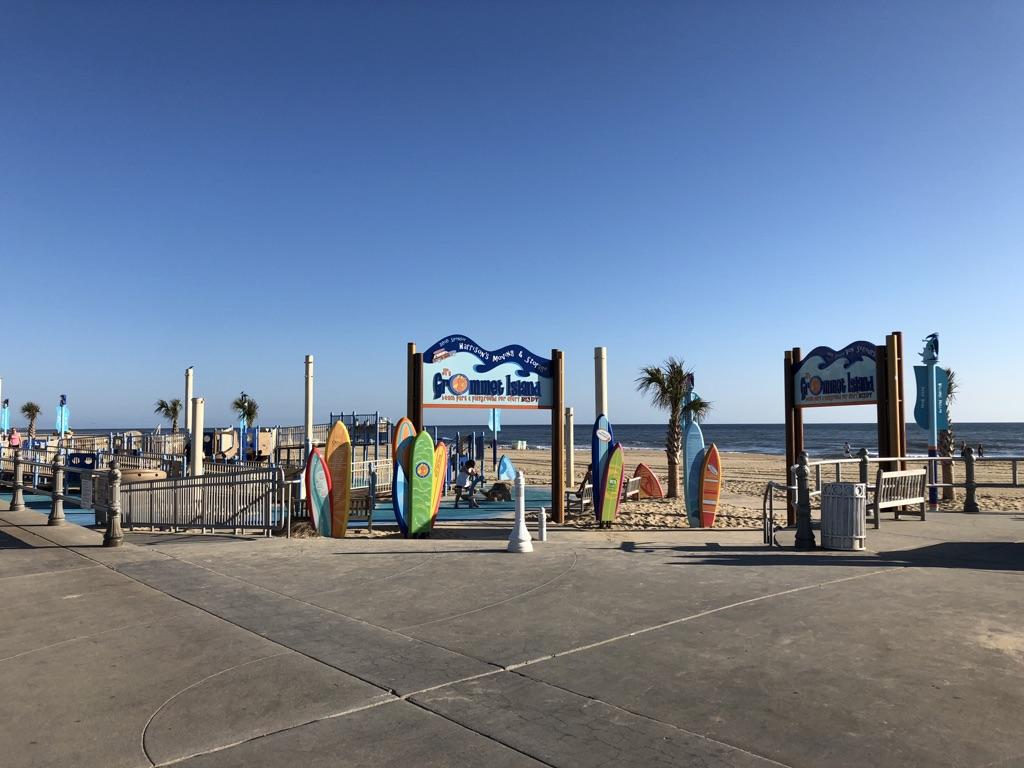 Colorful surfboards and signs on a boardwalk leading to a beach on a sunny day.