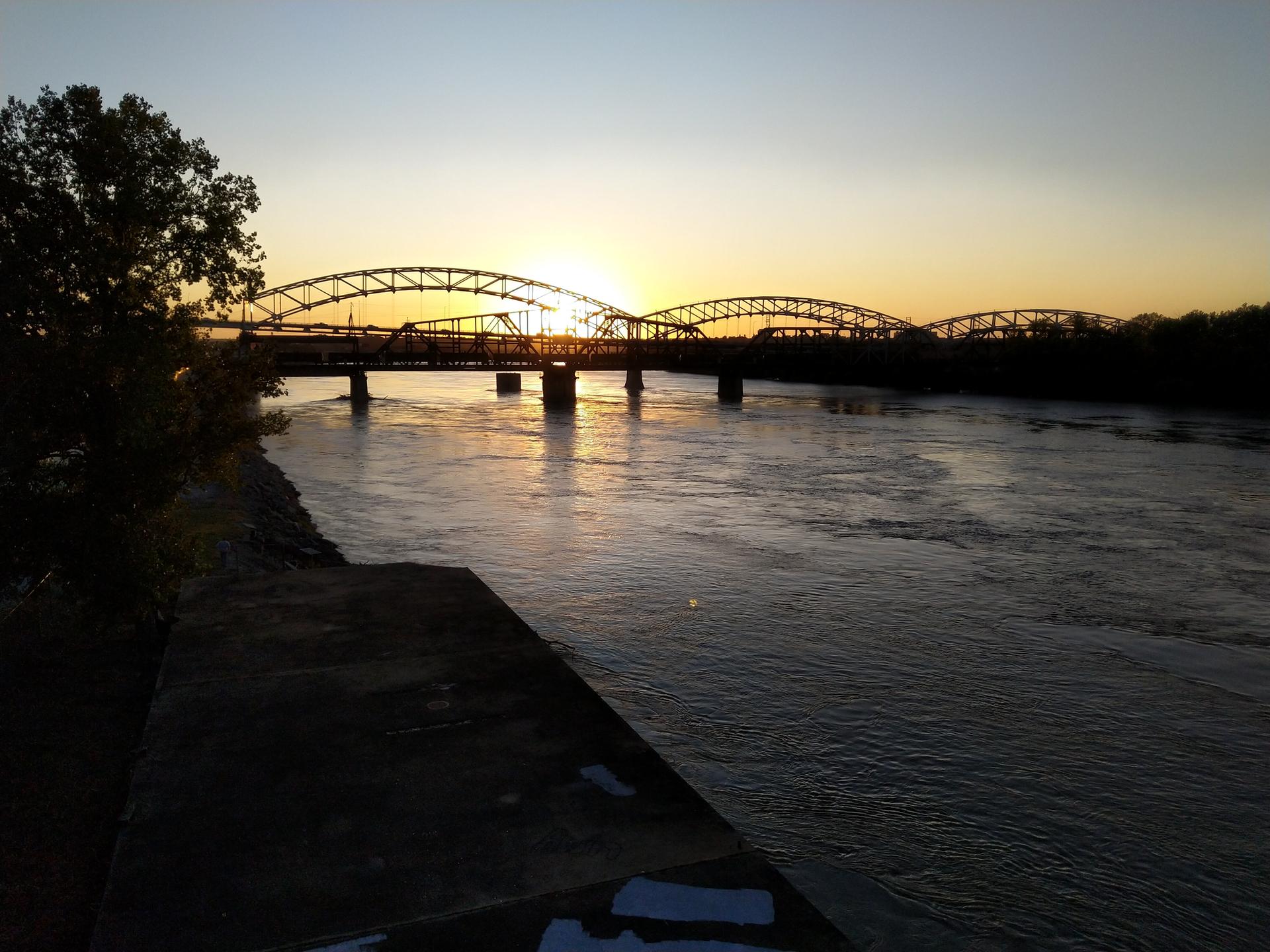 Arched bridges are silhouetted against a vibrant sunset over a wide river.
