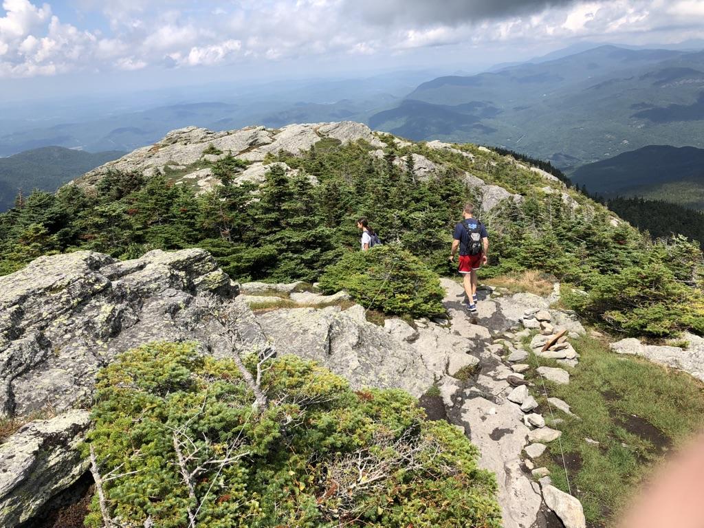 Two hikers on a rocky mountain ridge with evergreen trees under cloudy sky.