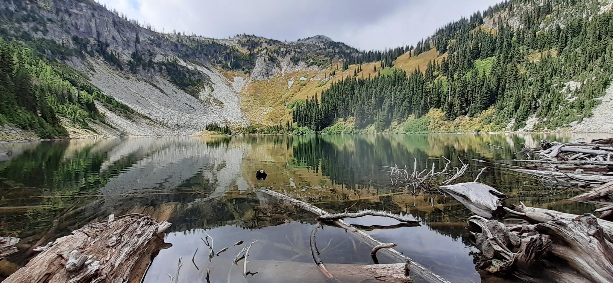 Calm mountain lake reflecting steep slopes and evergreen forests under cloudy sky.