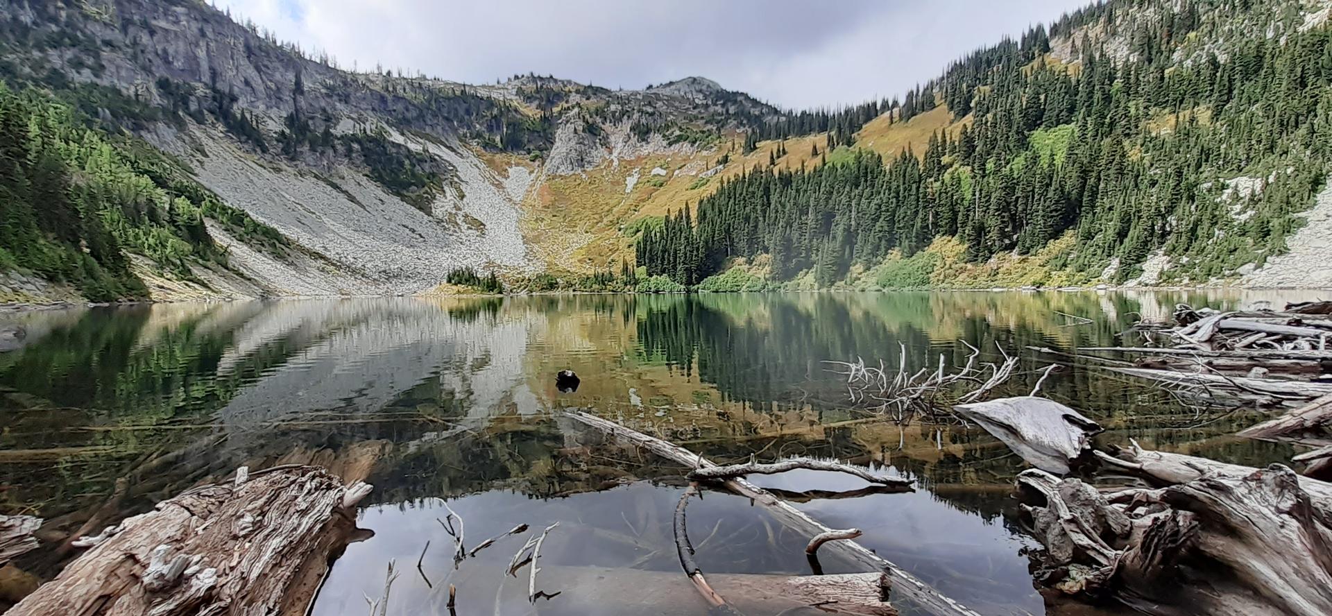 Calm mountain lake reflecting steep slopes and evergreen forests under cloudy sky.
