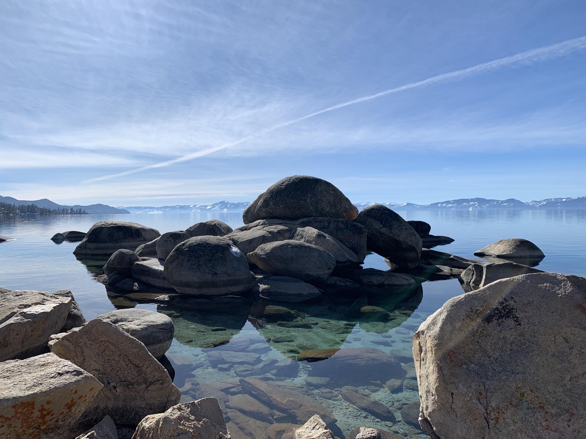 Large boulders reflected in clear, calm lake water under a bright sky.