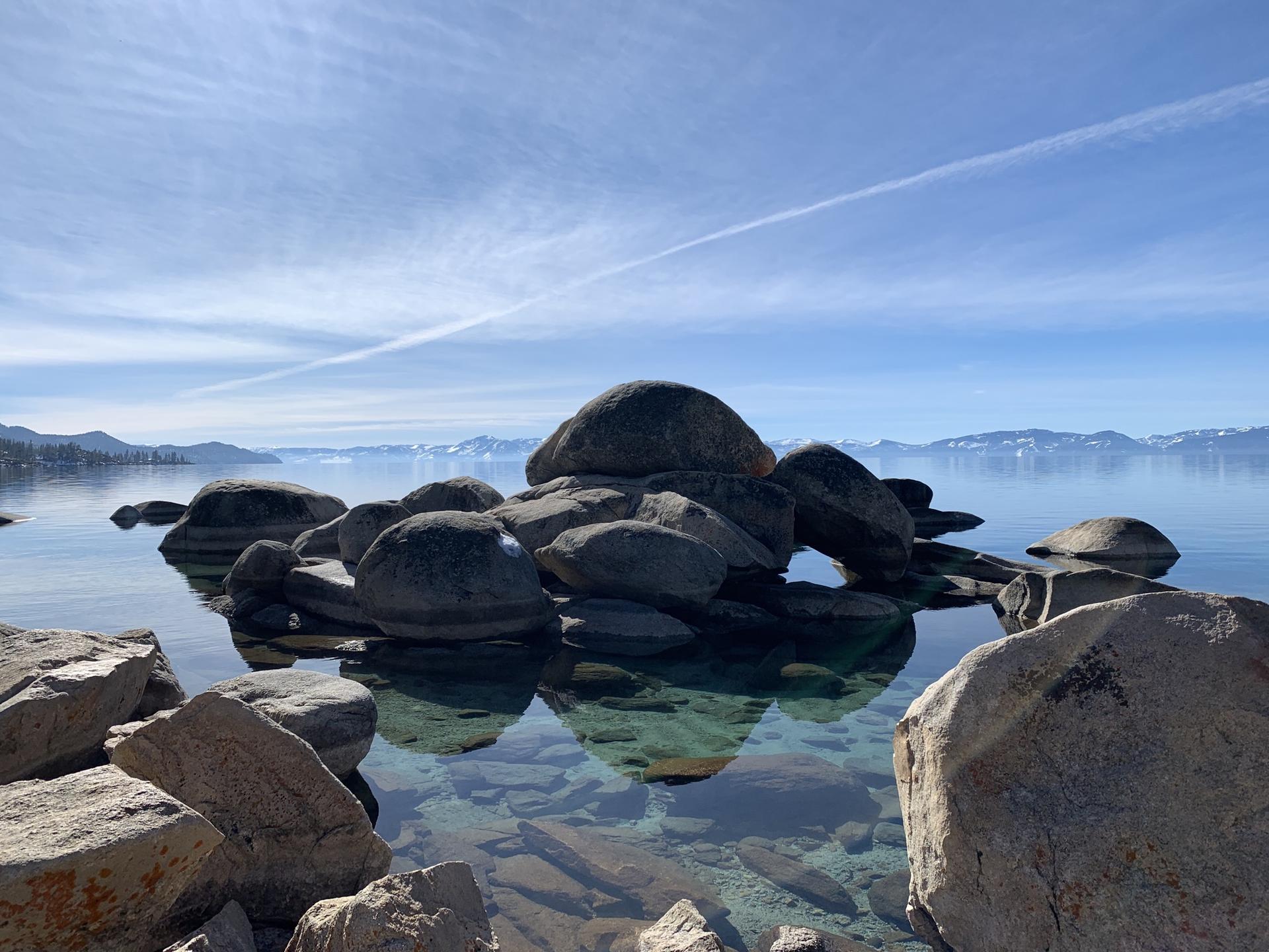 Large boulders reflected in clear, calm lake water under a bright sky.