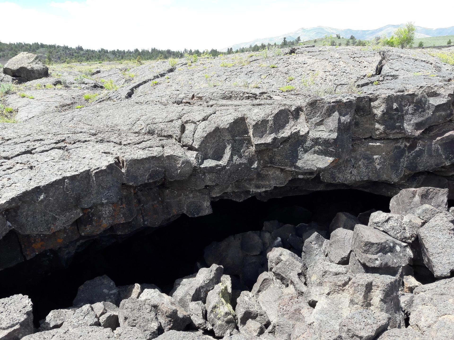 Dark cave opening in a rocky, volcanic landscape under bright daylight.