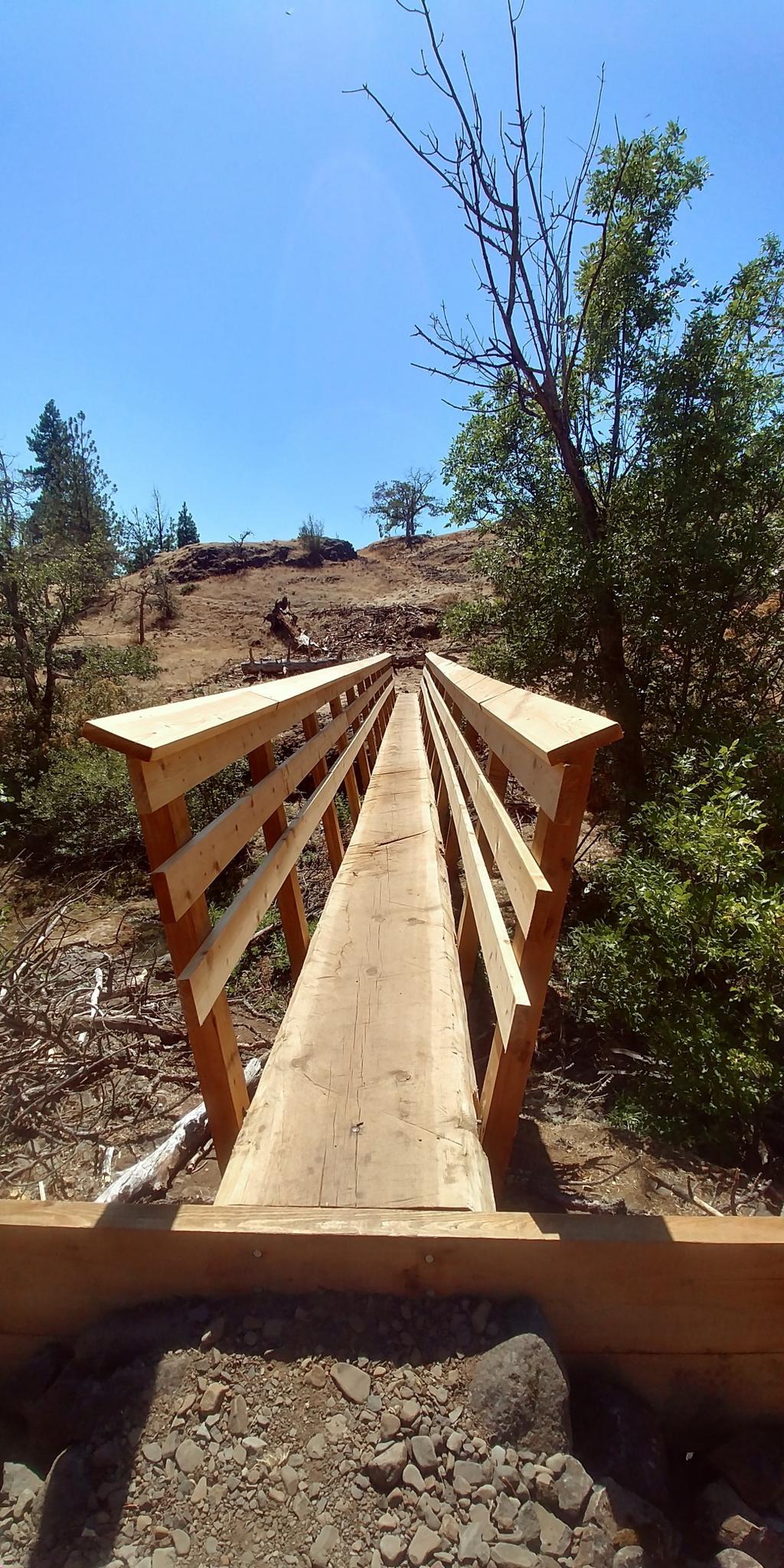 A new wooden footbridge spans a dry, rocky gully on a sunny day.
