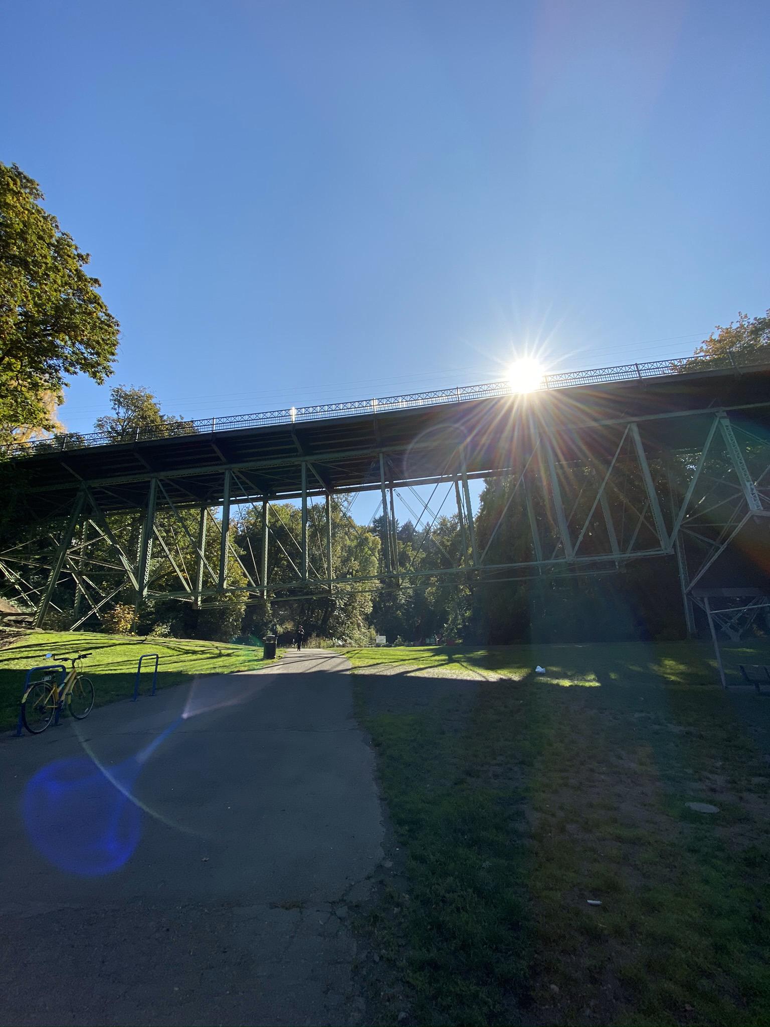 Sunburst over a large metal bridge spanning a path with parked bicycles.
