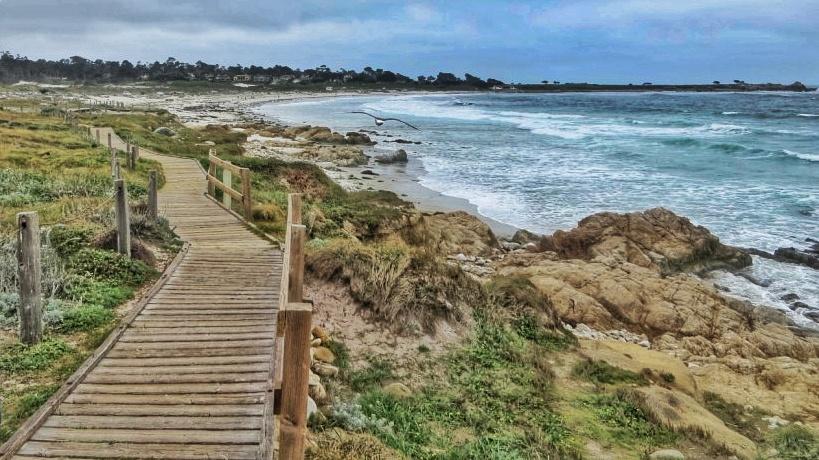 Wooden boardwalk along a rocky coastline with crashing waves under cloudy sky.