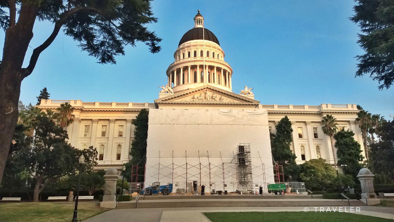 Grand domed building partially covered by scaffolding, surrounded by trees under clear sky.