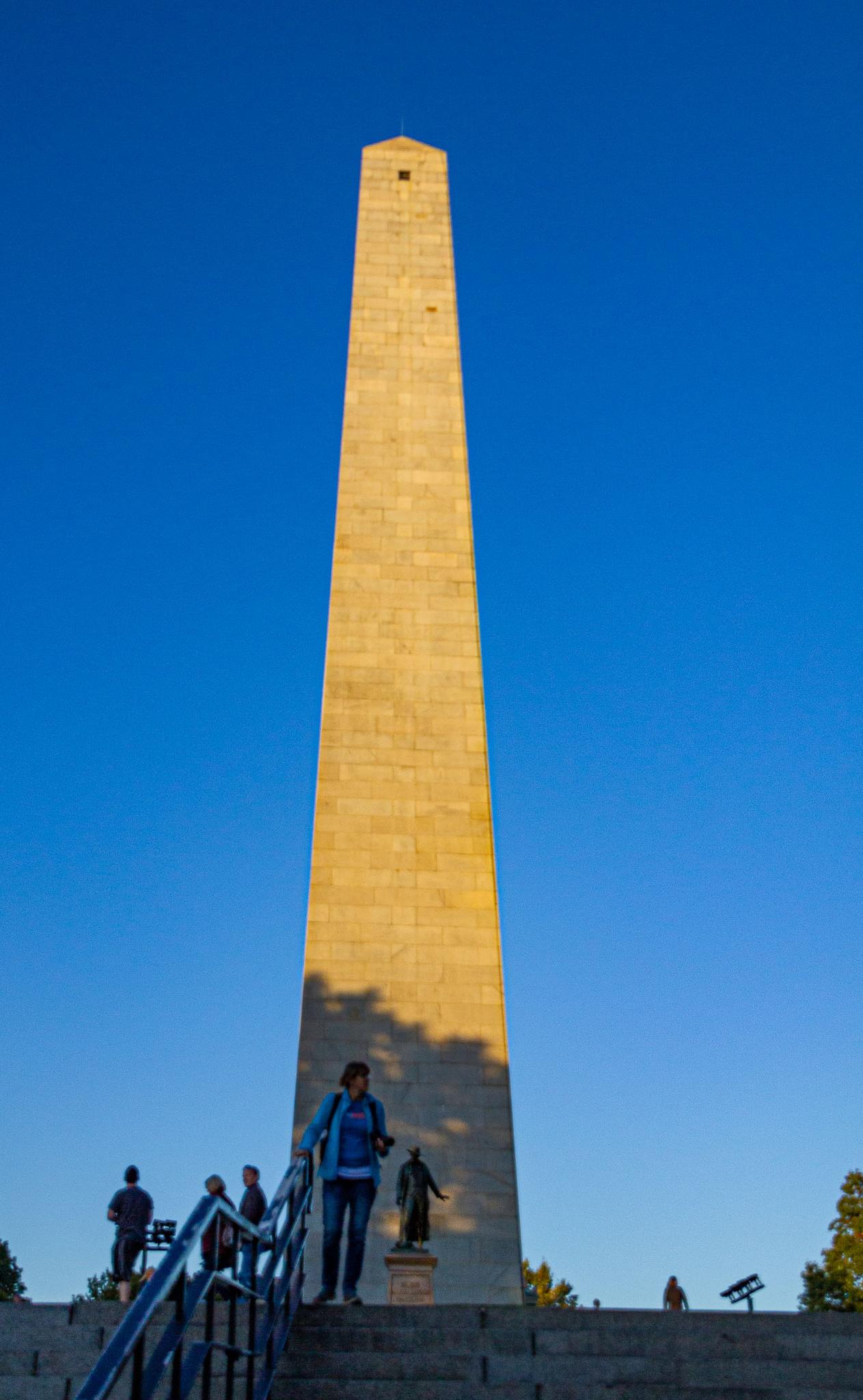 A tall stone obelisk stands against a clear blue sky, people on steps.