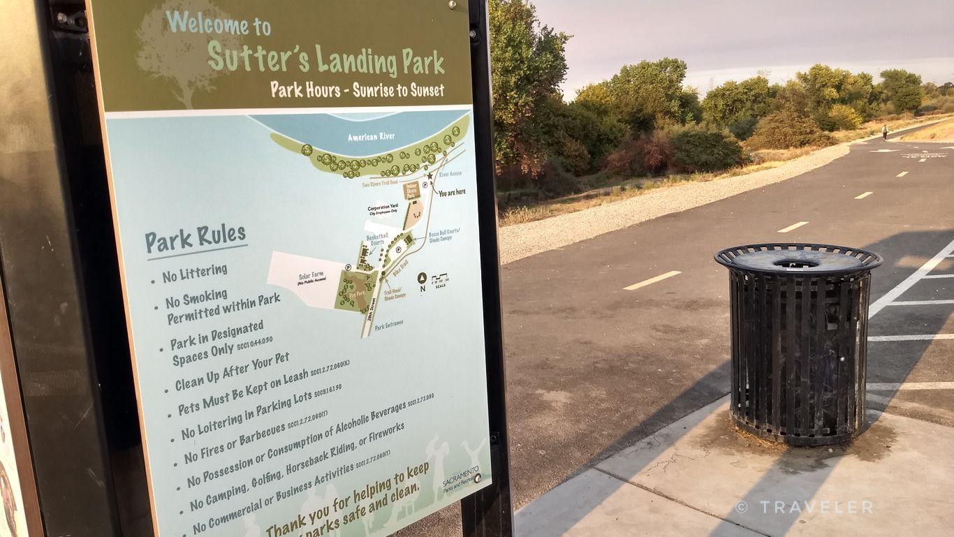 A park sign with rules and map next to a trash can and road.