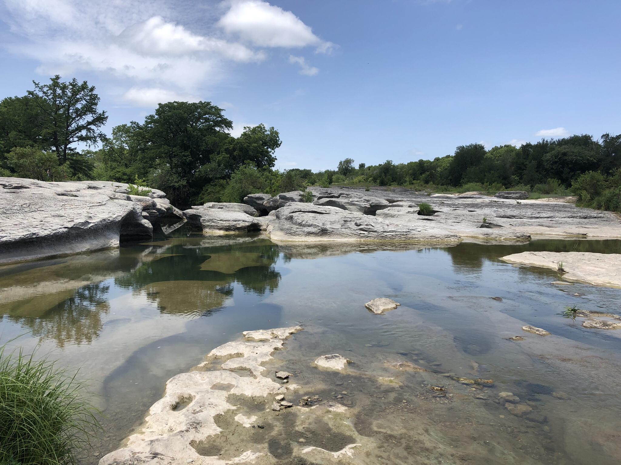 Wide river with rocky shallows, reflections, and green trees under blue sky.