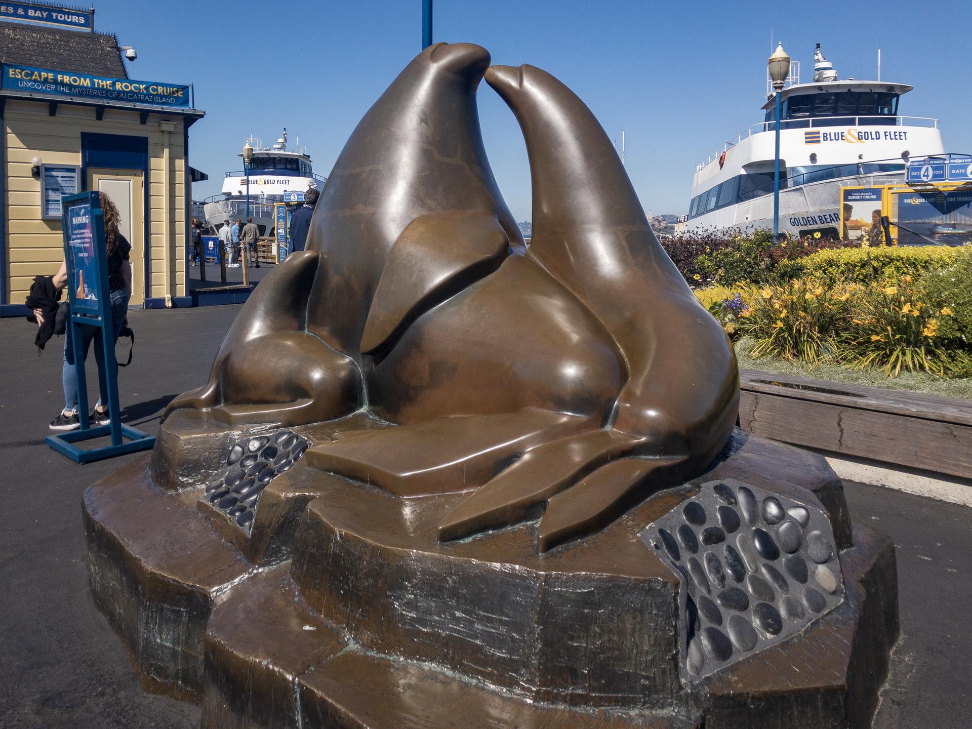 Bronze seal statues on a pier with boats and people nearby.