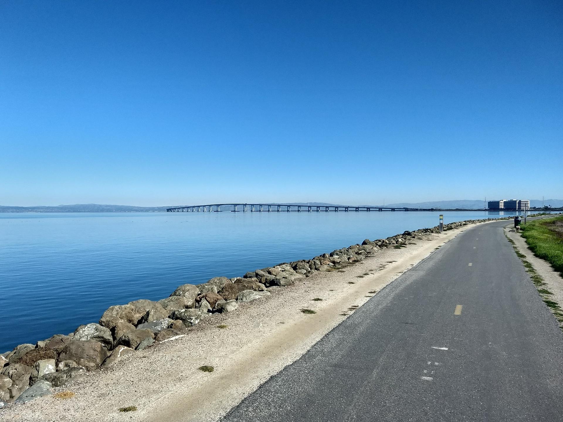 Paved path next to a rocky shoreline, calm blue water, and a distant bridge.