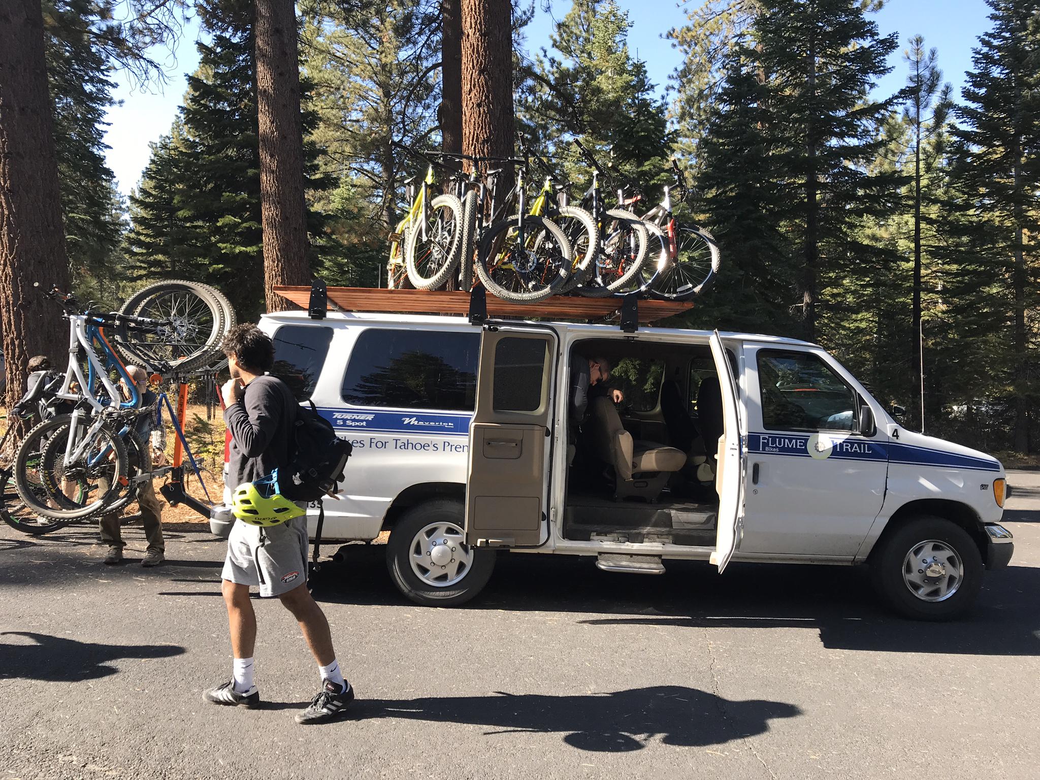 People loading bicycles onto a white van with a roof rack in a forest.