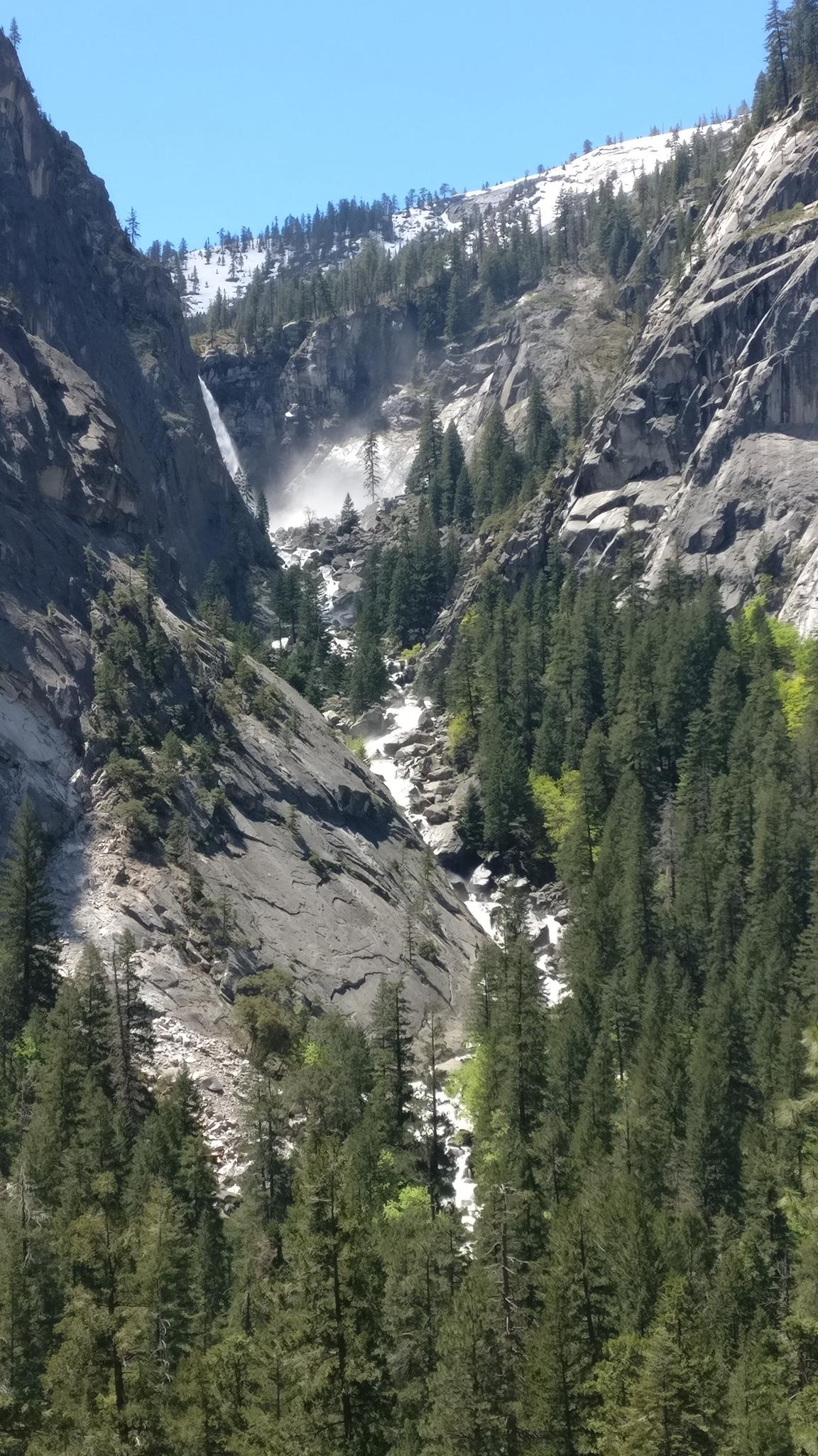 Distant waterfall cascades down a steep, forested mountain valley under a clear sky.