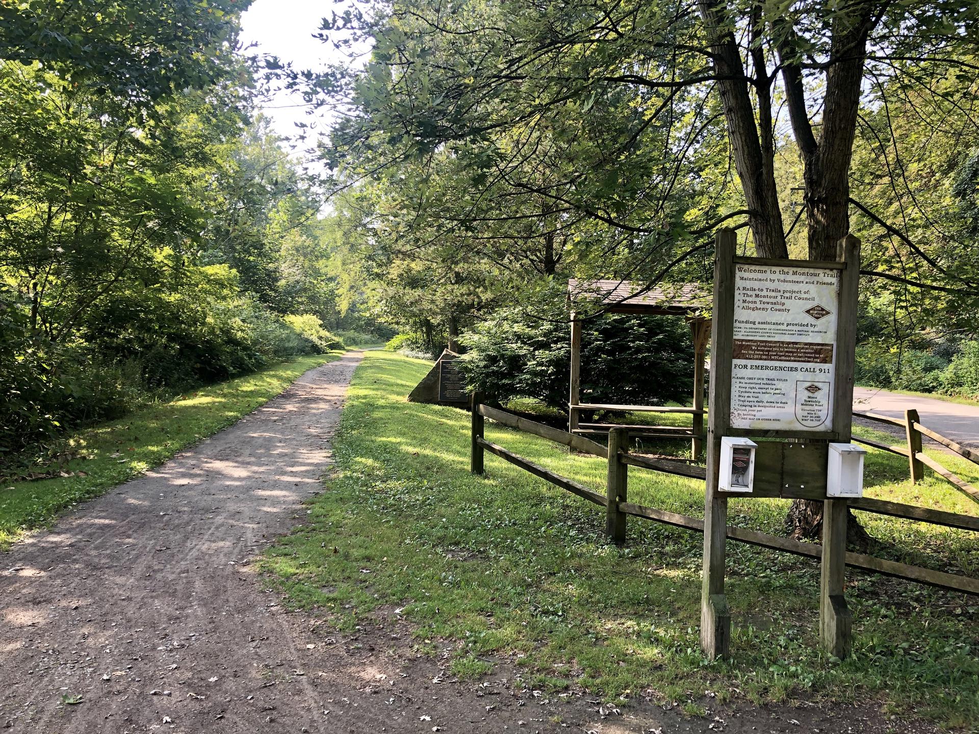 A sunny dirt path winds through green woods with an informational sign.