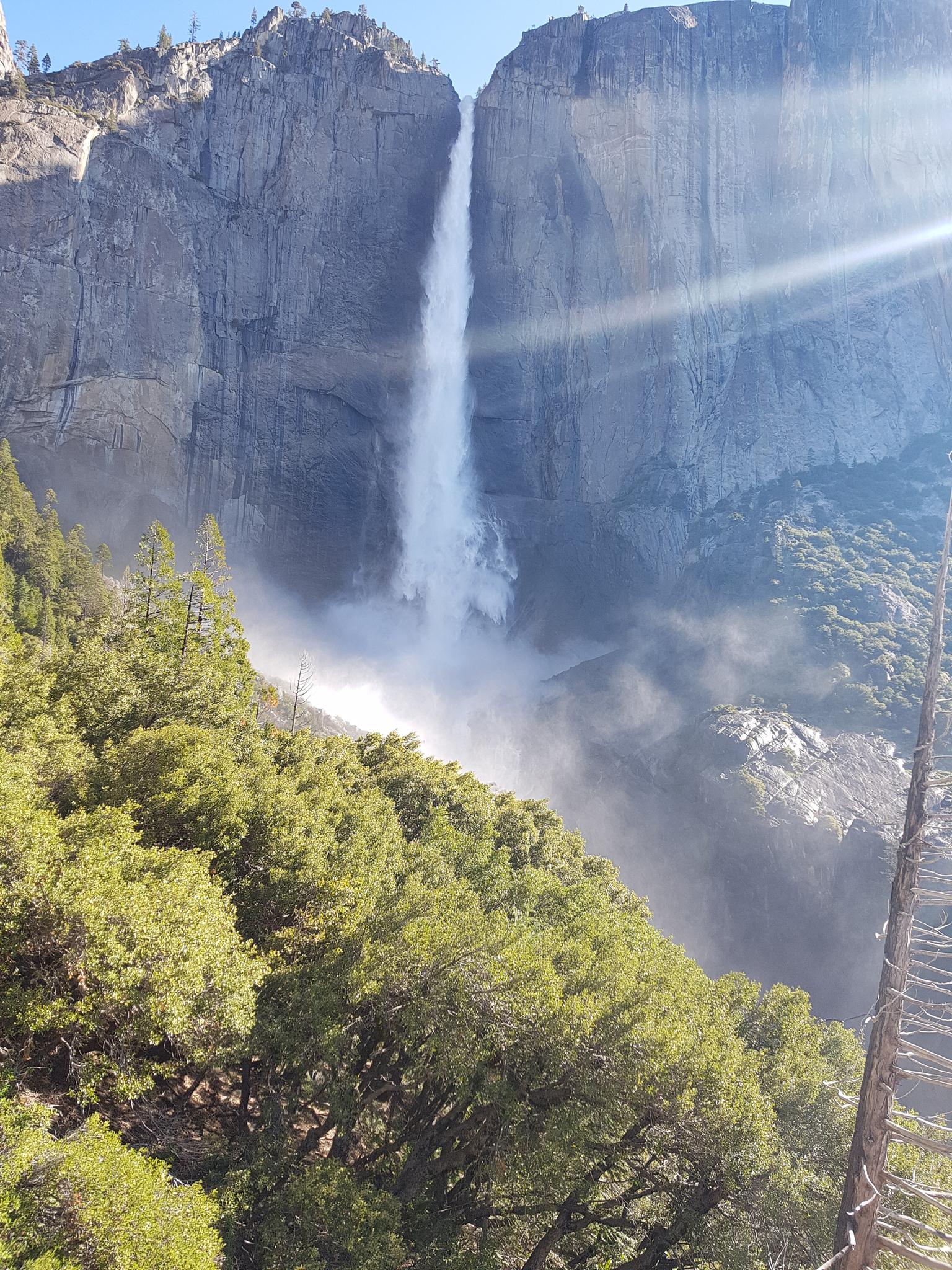 Tall waterfall cascades down rocky cliffs with bright sunlight and green trees.