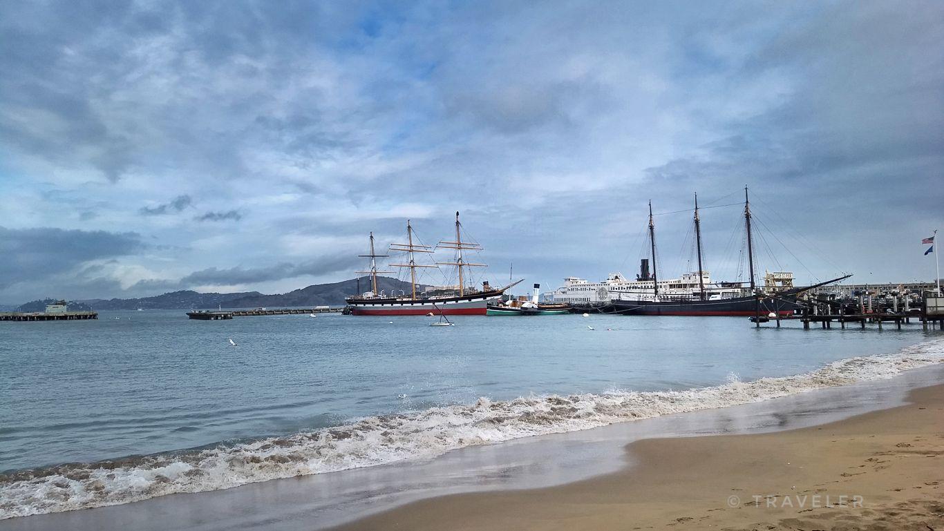 Tall ships docked in a bay, sandy beach with waves, cloudy sky.