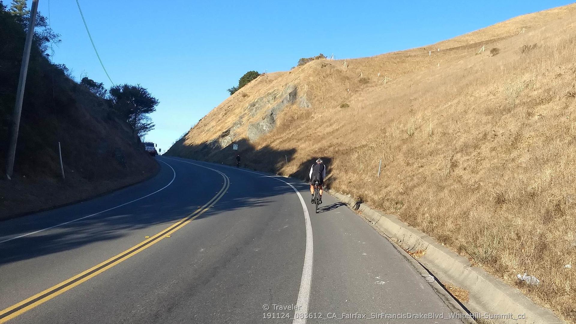 A cyclist rides on a sunny paved road beside a dry, golden hillside.