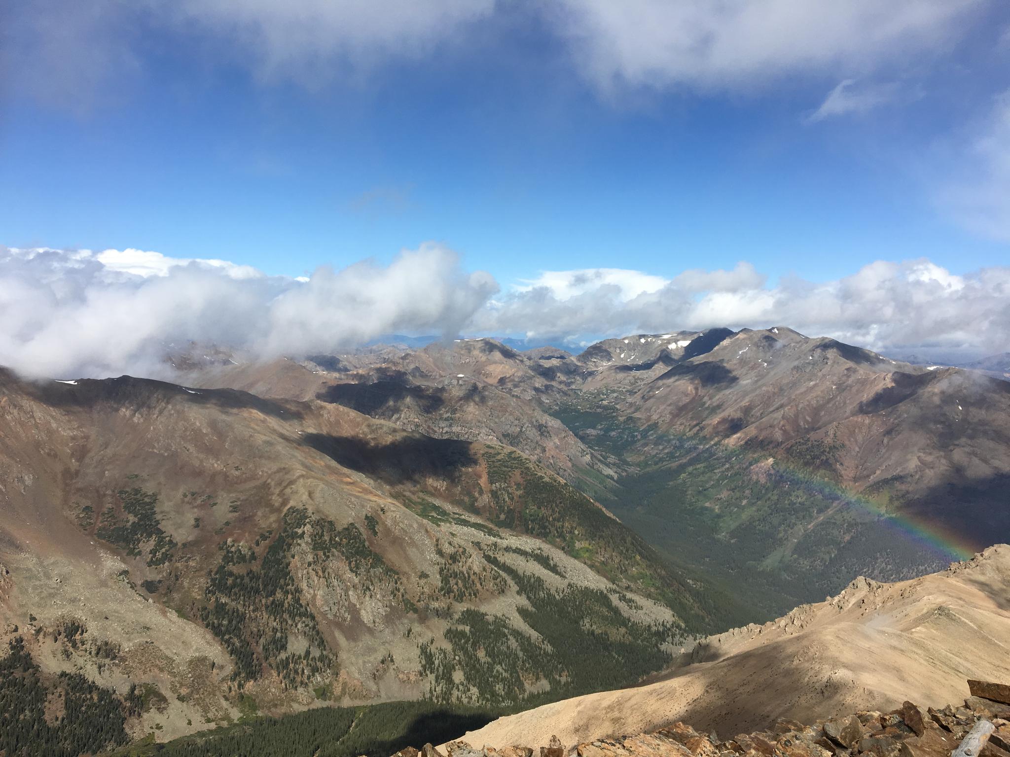 Expansive mountain landscape with clouds, a valley, and a faint rainbow.