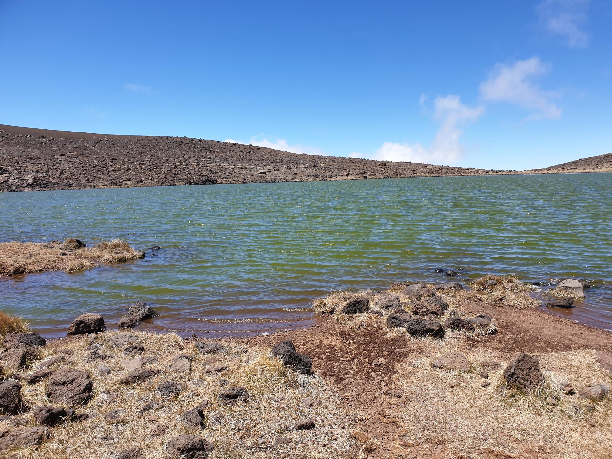Green lake with rocky shore, brown hills, and blue sky with clouds.
