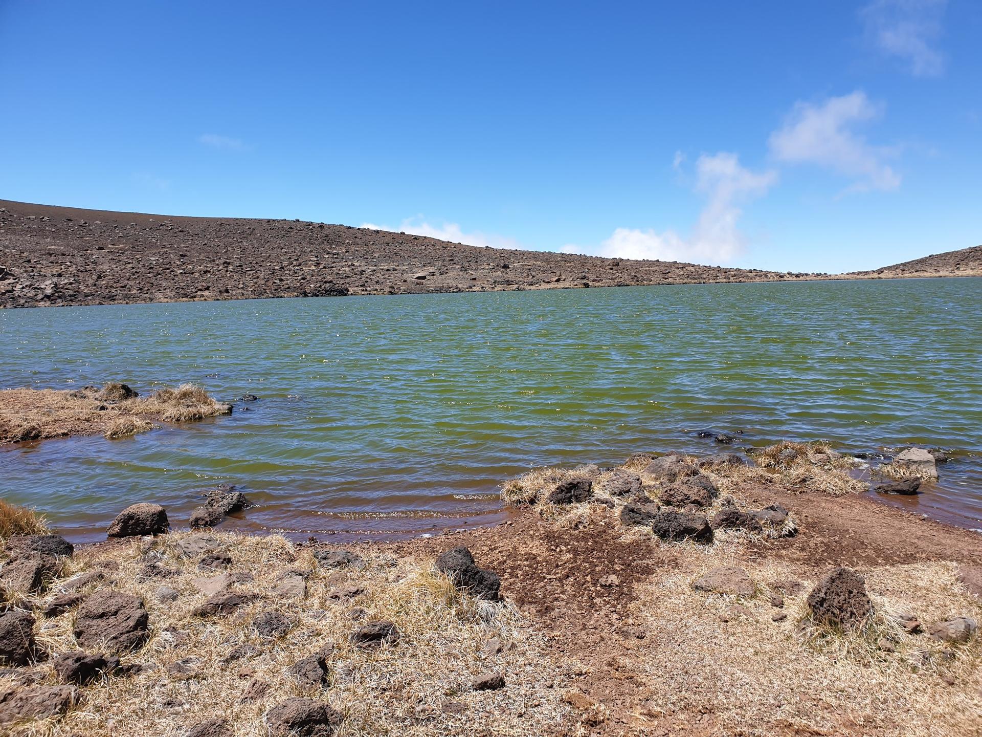 Green lake with rocky shore, brown hills, and blue sky with clouds.