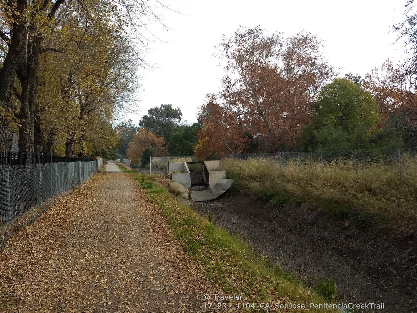 Leaf-covered dirt path beside a fence and concrete drainage under an overcast sky. - Penitencia Creek Park Loop