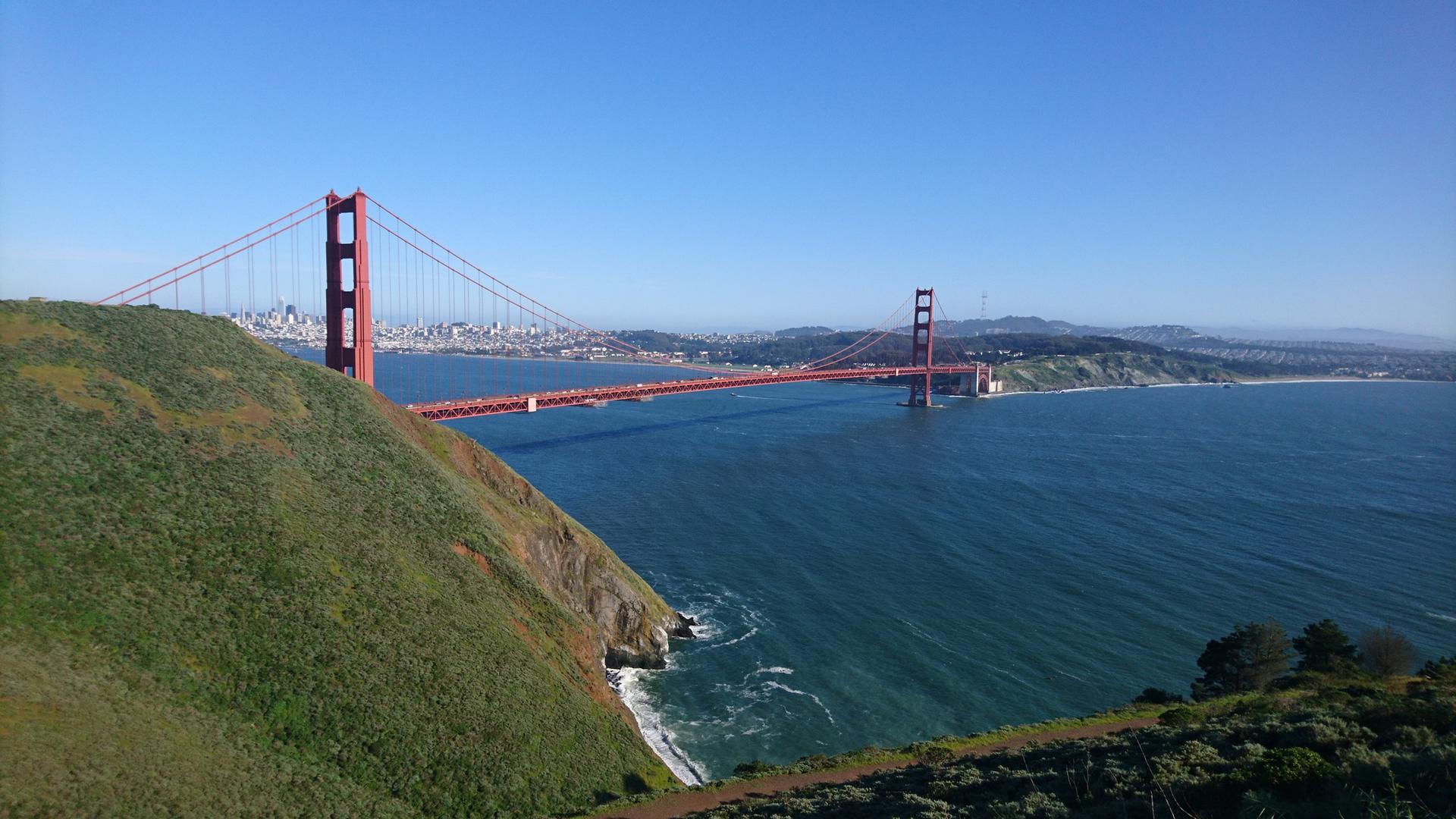 Red suspension bridge spanning a bay, with a steep green hillside. Sunny.