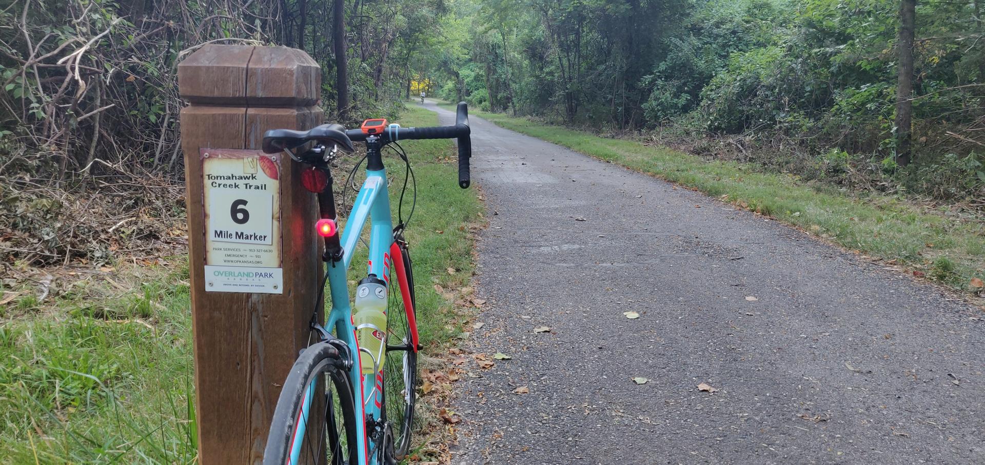 Bicycle leaning against a mile marker on a paved trail through green woods.