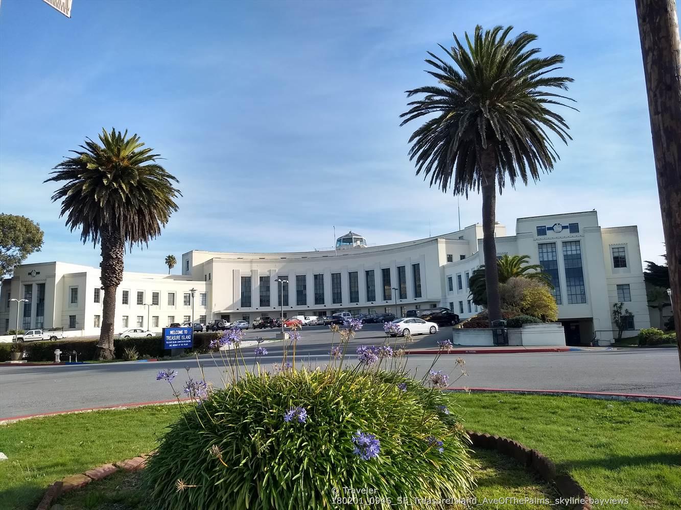 Large white building with palm trees and a circular flower bed in front. Bright day.